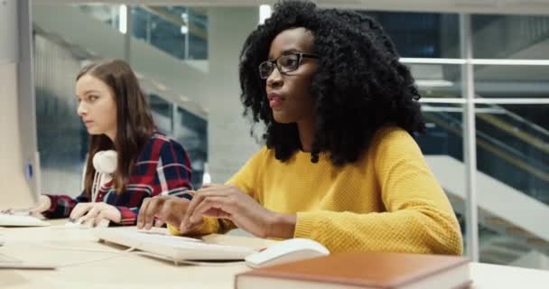 Two smart girls students sit in information room and solve tests on computers. African American girl with curly hair gives advice to friend. — Stock Video