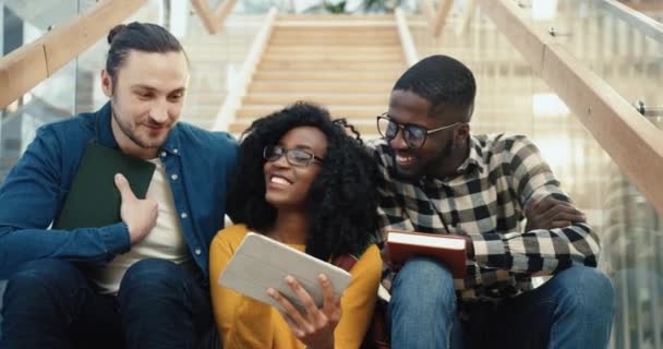 Tres estudiantes multinacionales se sientan juntos en las escaleras universitarias durante el descanso y ven un interesante video en la tableta y hablando. — Vídeo de stock