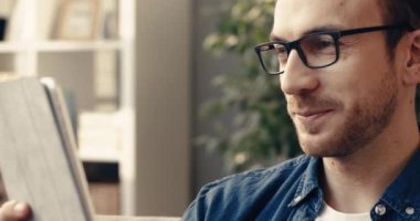 Gözlüklü yakışıklı beyaz adamın evdeki kanepede oturup arkadaşlarıyla video yoluyla iletişim kurmak için tablet kullandığı yakın plan..