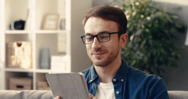 Gözlüklü yakışıklı sakallı adamın yakın plan fotoğrafı evdeki koltukta oturuyor ve tablet kullanarak sosyal ağlardan arkadaşlarıyla iletişim kuruyor..