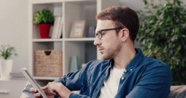 Genç adam kanepeye oturur ve tabletine çabucak mesaj yazar, sonra başını çevirir ve kameraya bakar. Başarılı bir adamın portresi.