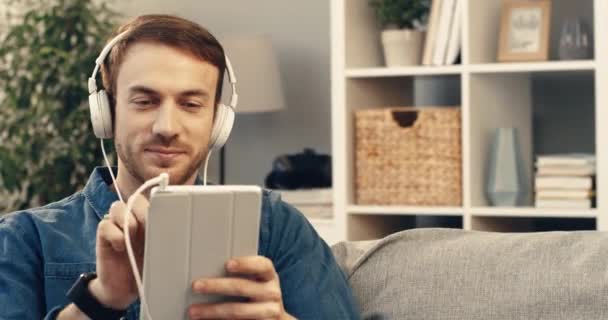 Young happy bearded man in denim shirt sits on couch at home and listening music on headphones using tablet. — Stock Video