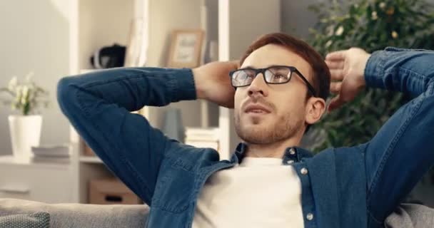 Joven barbudo con camisa de mezclilla y gafas se sienta en casa en el sofá y descansa con las manos detrás de la cabeza. — Vídeo de stock