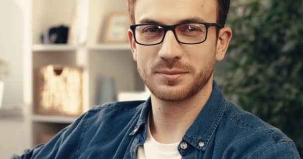 Close-up retrato de homem barbudo bonito com óculos sentados em casa no sofá e olhando para a câmera sorrindo. — Vídeo de Stock