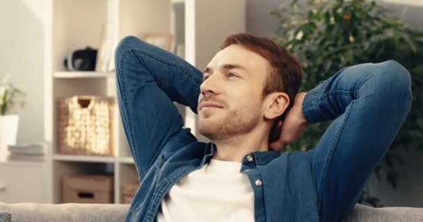 Jovem barbudo de camisa jeans senta-se em casa no sofá e descansa com as mãos atrás da cabeça sonhando com fim de semana. — Vídeo de Stock