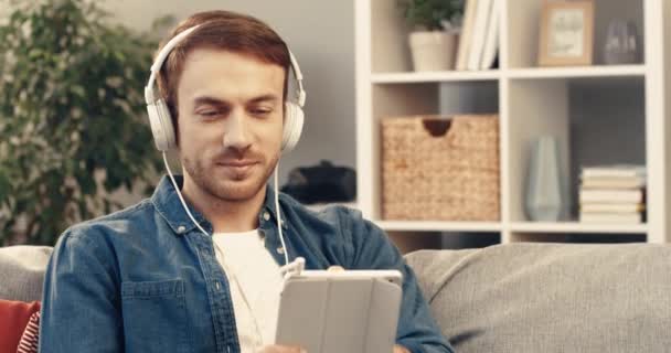Ung vit man sitter hemma på soffan i vita hörlurar och lyssnar musik på sin surfplatta njuter med bra ljud. — Stockvideo