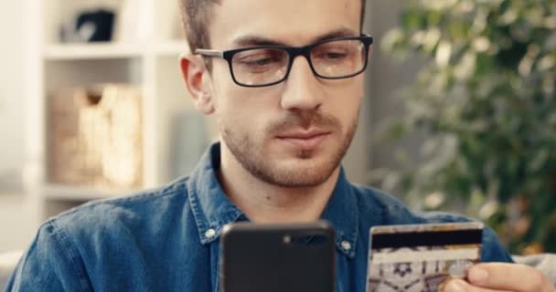 Κοντινό πλάνο του Καυκάσου με γυαλιά που κρατάει πιστωτική κάρτα και smartphone ενώ κάθεται στον καναπέ στο σπίτι και απολαμβάνει την επιτυχή αγορά. — Αρχείο Βίντεο