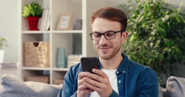 Joven hombre caucásico en gafas se sienta en casa y escribiendo mensaje a su novia mientras sostiene el teléfono inteligente y sonríe. — Vídeos de Stock