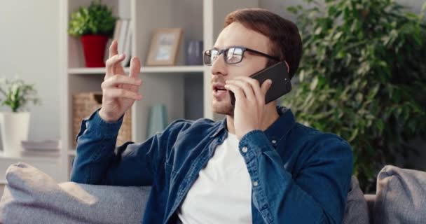 Adulto bell'uomo caucasico con gli occhiali si siede sul divano a casa e parla al telefono con il suo migliore amico. — Video Stock