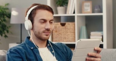 Kot tişörtlü, mutlu sakallı genç adam evdeki koltukta oturup kulaklıkla tablette film izliyor. .