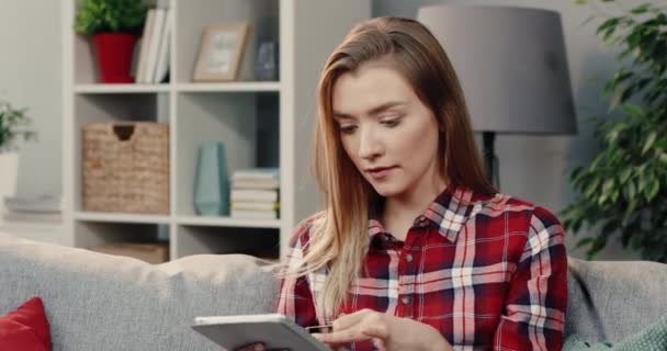 Jeune belle blonde assise à la maison sur le canapé avec tablette et carte bancaire et fait des achats en ligne. — Video