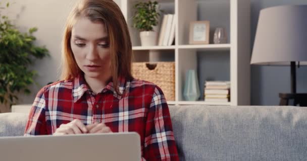 Wanita muda berambut pirang Kaukasia yang duduk sendirian di sofa rumah dan fokus untuk mengerjakan laptop. — Stok Video