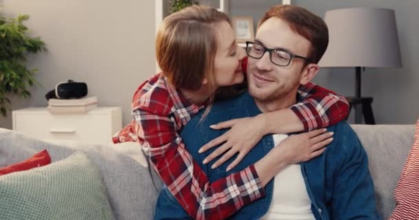 Retrato de una joven feliz pareja casada sentada en casa en el sofá y mirando a la cámara sonriendo. Esposa abraza hombre por cuello y lo besa en la mejilla. — Vídeos de Stock