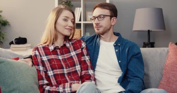 Νεαρό ευτυχισμένο ζευγάρι Καυκάσιος κάθεται στο σπίτι στον καναπέ γυρίζοντας το κεφάλι τους και κοιτάζοντας κάμερα χαμογελώντας. — Αρχείο Βίντεο