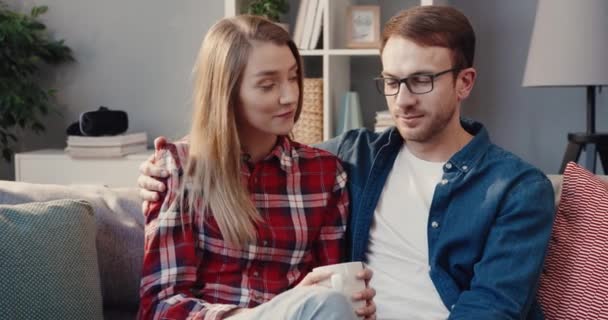 Крупним планом молода щаслива пара сидить вдома на дивані і проводить вільний час разом розмовляючи і п'ючи гарячим чаєм . — стокове відео