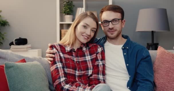 รูปภาพของคู่แต่งงานสาวที่มีความสุขนั่งที่บ้านบนโซฟายิ้มที่กล้อง ผู้ชายกอดภรรยาบนไหล่ . — วีดีโอสต็อก