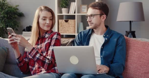 Genç evli çift evlerinde kanepede birlikte vakit geçiriyorlar. Kocası dizüstü bilgisayarda çalışıyor, karısı akıllı telefon kullanıyor.. — Stok video