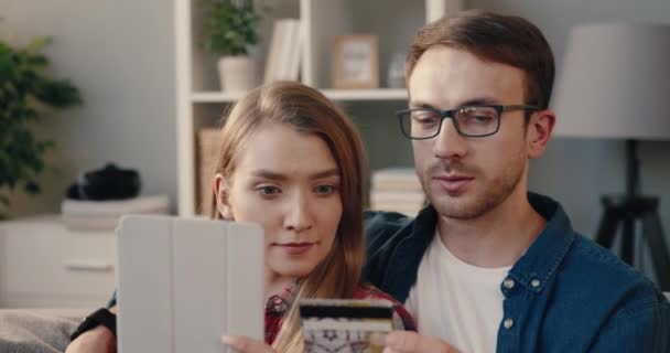 Unga glada par sitter på soffan med bärbar dator och gör online-överföring genom att ange bankkortsnummer. — Stockvideo