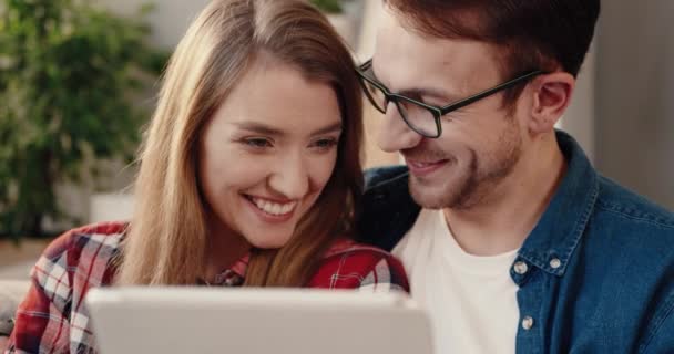 Close up of beautiful young couple sitting at home on couch and looking at funny video on tablet . — Stock Video