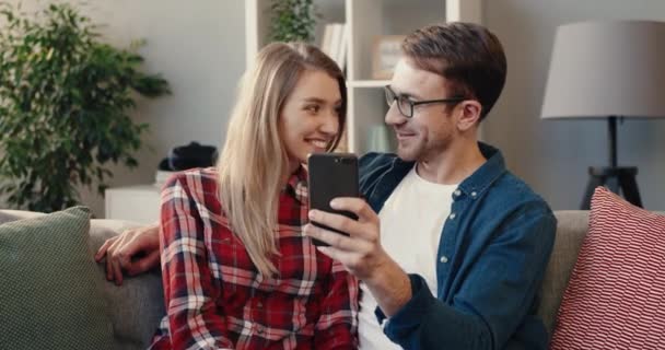 El marido feliz muestra la foto por teléfono a su mujer sentada en casa en el sofá. Joven pareja casada pasando tiempo libre juntos. — Vídeos de Stock