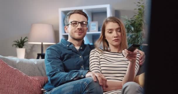 Fiatal pár otthon ül a kanapén és filmet néznek. Két ember együtt tölti a szabadidejét. A nő folyamatosan csatornát vált a távirányítóval. — Stock videók