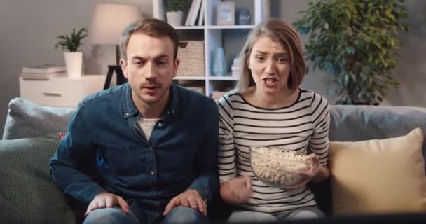 Young couple sitting at home on couch with popcorn and watching match on TV cheering for their favorite team. — Stock Video
