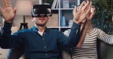 Genç evli çift evlerinde kanepede oturuyor ve sanal gerçeklik gözlükleriyle video oyunu oynuyorlar..