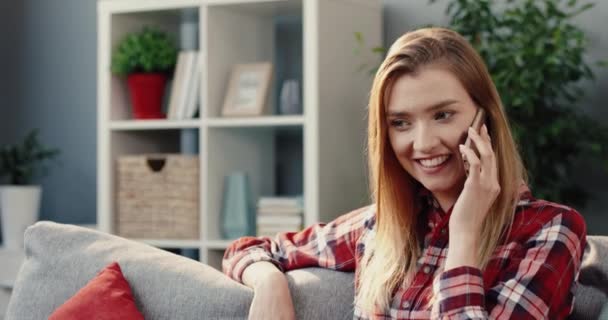Ung vacker blond flicka sitter hemma på soffan och pratar med vän på mobilen diskuterar nytt skvaller. — Stockvideo