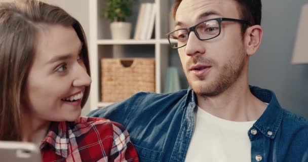 Jovem feliz casal caucasiano sentado em casa no sofá e se divertindo assistindo vídeo online usando smartphone. — Vídeo de Stock