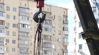 Mobil bir kule vincinin kancası, inşaat malzemelerini mavi bir gökyüzünün arka planına ve balkonları ve kliması olan çok katlı bir binaya karşı demir iplerle kaldırır. Kapat..