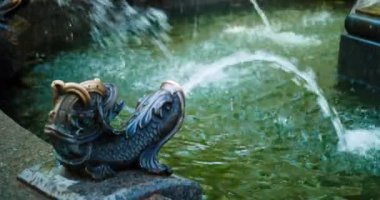 Şehir parkında modern tarzda güzel bir çeşme. Mavi elbiseli, çiçekli ve kahverengi çantalı bir kadın muhteşem bir bakır balığının sularının altında ellerini yıkıyor. Su sıçraması. Kapat.