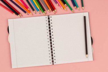 Açık okul karesi defter ve pembe arka planda renkli kalemler. Masanın üzerinde boş beyaz bir sayfa kağıt var. Ofis malzemeleri masaüstünde. Okula dönelim. Uzayı metin için kopyala. Üst görünüm