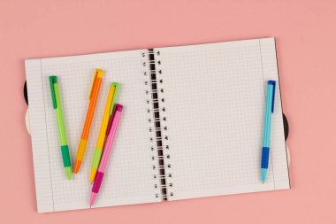 Açık okul karesi defter ve pembe arka planda renkli kalemler. Masanın üzerinde boş beyaz bir sayfa kağıt var. Ofis malzemeleri masaüstünde. Okula dönelim. Uzayı metin için kopyala. Üst görünüm