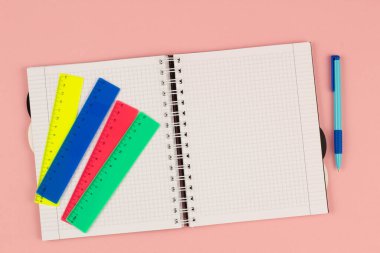 Açık okul karesi defter, pembe arka planda kalem ve renk cetvelleri. Masanın üzerinde boş beyaz bir sayfa kağıt var. Ofis malzemeleri masaüstünde. Okula dönelim. Uzayı metin için kopyala. Üst görünüm