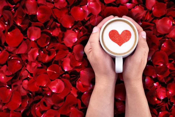 Hombre sosteniendo taza caliente de leche sobre fondo de pétalos de rosa roja, con forma de corazón rojo — Foto de Stock