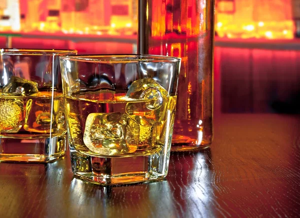 Vasos de whisky con hielo en la mesa de bar cerca de la botella de whisky en un ambiente cálido — Foto de Stock