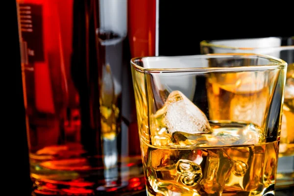 Close-up of whiskey with ice in glasses near bottle on black background — Stock Photo, Image