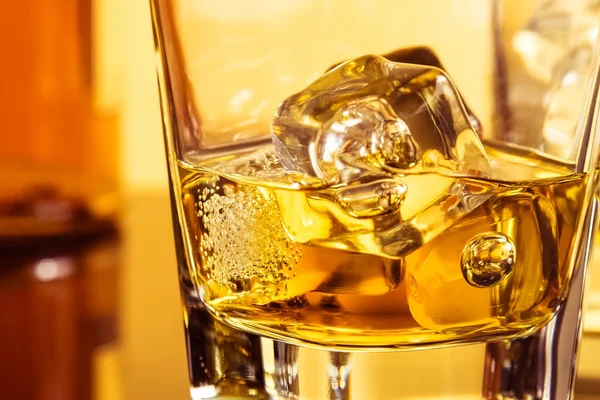 Dettaglio di bicchiere di whisky con ghiaccio vicino bottiglia sul tavolo con riflesso, atmosfera calda — Foto Stock