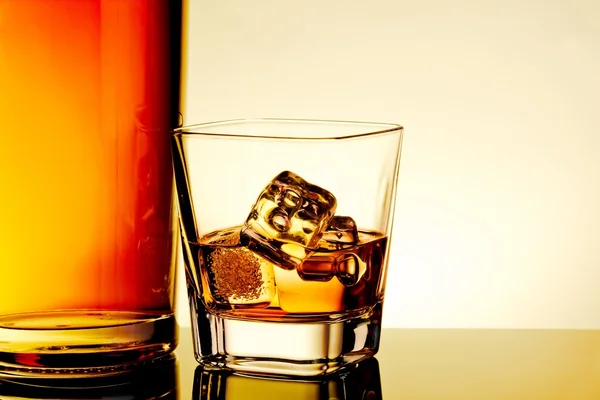 Glass of whiskey with ice cubes near bottle on table with reflection, warm tint atmosphere — Stock Photo, Image