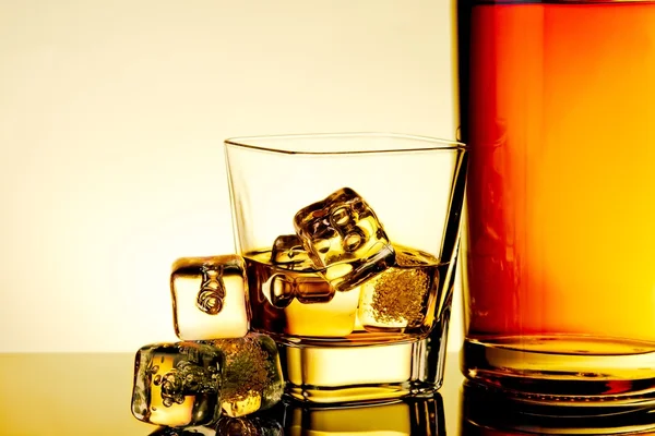 Glass of whiskey with ice cubes near bottle and ice cubes on table with reflection, warm tint atmosphere — Stock Photo, Image