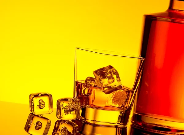 Glass of whiskey with ice cubes near bottle on table with reflection, warm yellow tint atmosphere — Stock Photo, Image