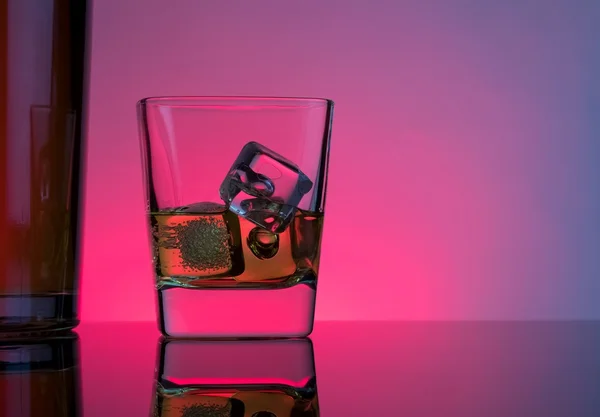 one glass of whiskey with ice cubes near bottle on table with reflection, lights disco atmosphere