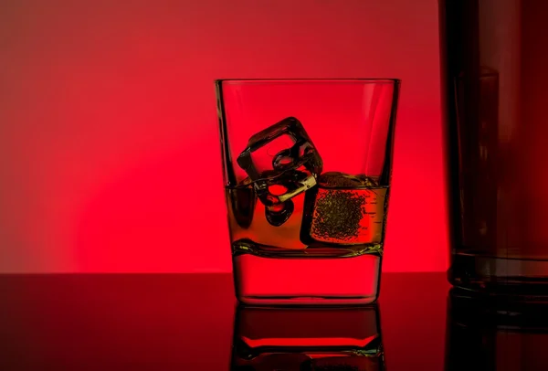 Un vaso de whisky con cubitos de hielo cerca de la botella en la mesa con reflexión, luces rojas ambiente disco — Foto de Stock