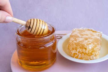 a woman's hand takes honey on a wooden stick from a full jar of honey with honeycombs on a plate. dipper takes honey from a bowl full with space for your text