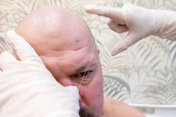 Men threading is the removal of unwanted vegetation on the body with a thread. a female cosmetologist plucks a man's eyebrows with a thread