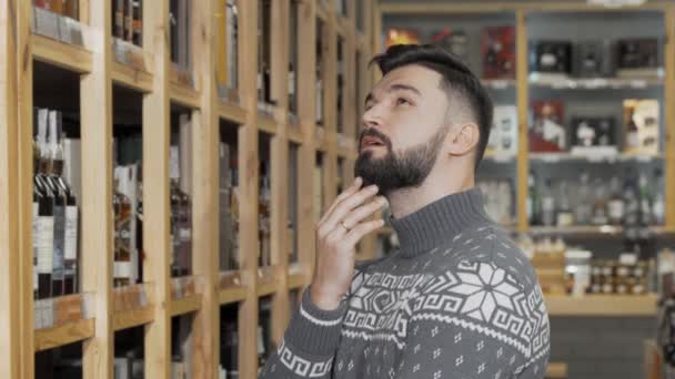Bonito homem barbudo olhando para as prateleiras com garrafas na loja de vinhos — Vídeo de Stock
