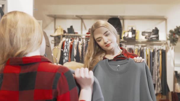 Νεαρή γυναίκα εξετάζει ρούχα μπροστά από τον καθρέφτη στο κατάστημα μόδας — Αρχείο Βίντεο