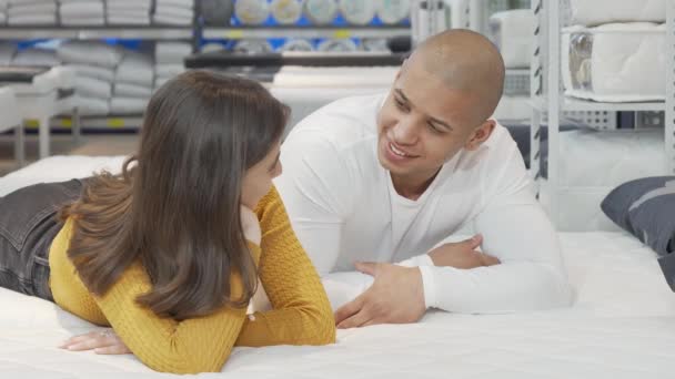 Pareja joven hablando mientras está acostada en una cama nueva en la tienda de muebles — Vídeo de stock