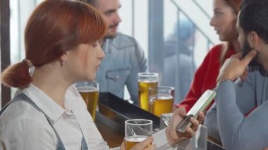 Barda bira içerken akıllı telefonunu kullanan genç bir kadın.