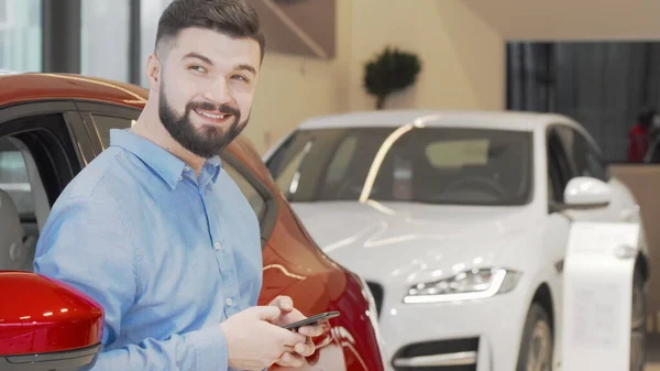 Ευτυχισμένος άνθρωπος γραπτών μηνυμάτων σε ένα έξυπνο τηλέφωνο, ενώ ψώνια για το νέο αυτοκίνητο στην αντιπροσωπεία Εικόνα Αρχείου